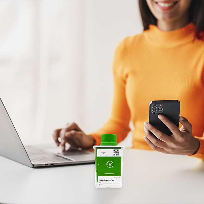 Person holding mobile phone with laptop and Saliva Test Cup